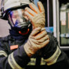 Feuerwehrmann mit Atemschutzmaske zieht Penkert Elk Defender 3.0 Handschuhe mit langem Stulpen an, bereit für den Einsatz in gefährlicher Umgebung.