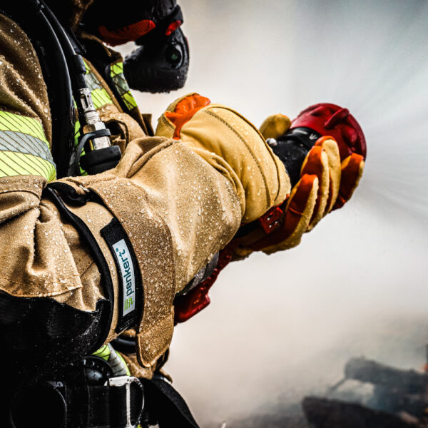 Feuerwehrmann im Einsatz trägt Penkert Elk Defender 3.0 Handschuhe mit langem Stulpen, hellbraunes Design mit orangen Akzenten, hält einen Feuerwehrschlauch bei einem Löscheinsatz.