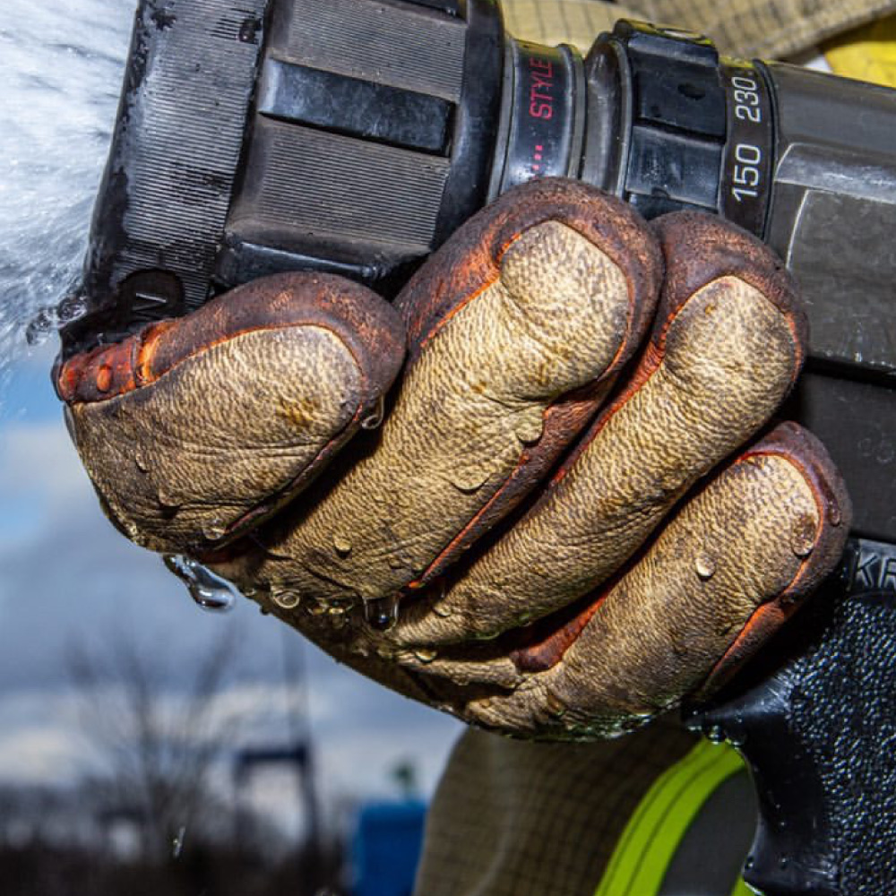 Ein Feuerwehrmann hält mit nassen Penkert Elk Defender 3.0 Kurzvariante Handschuhen einen Feuerwehrschlauch, während Wasser spritzt.