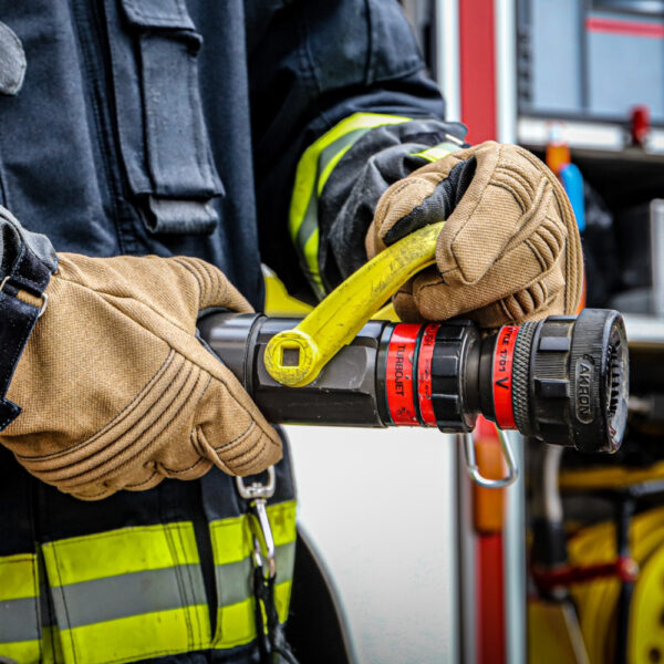 Ein Feuerwehrmann trägt Penkert FireDevil 911 X-treme kurze Handschuhe und bedient einen Feuerlöschschlauch, wobei die reflektierenden Streifen an der Schutzkleidung sichtbar sind.