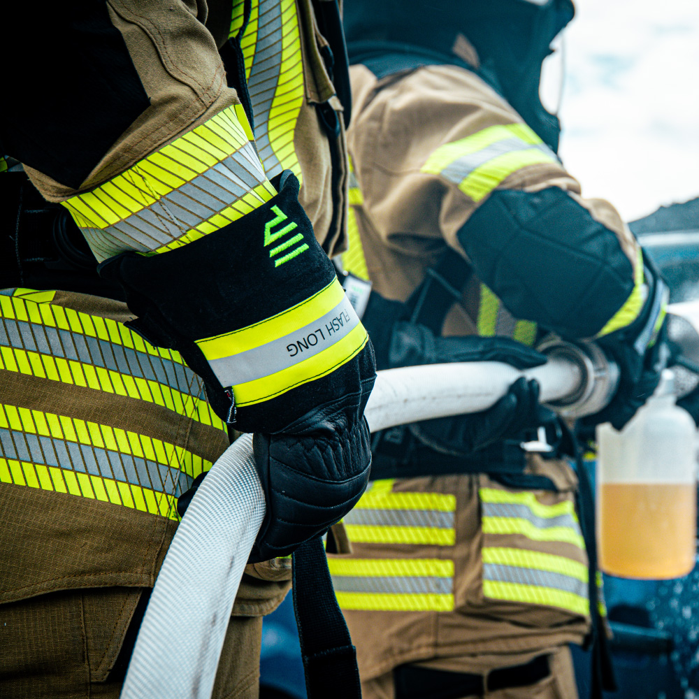 Das Bild zeigt zwei Feuerwehrleute im Einsatz, die einen Feuerwehrschlauch halten. Beide tragen vollständige Schutzkleidung mit reflektierenden gelben Streifen und schwarze Feuerwehrhandschuhe mit der Aufschrift "FLASH LONG". Der Fokus liegt auf den Handschuhen und dem Schlauch, während die Feuerwehrleute in Aktion sind. Die Szene vermittelt ein Gefühl von Professionalität und Einsatzbereitschaft.
