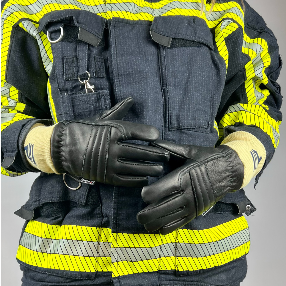 Das Bild zeigt einen Feuerwehrmann in Schutzkleidung mit reflektierenden gelben Streifen. Der Fokus liegt auf den schwarzen Feuerwehrhandschuhen mit gelben Manschetten, die der Feuerwehrmann trägt. Die Hände sind vor dem Körper verschränkt, was die Struktur und das Design der Handschuhe deutlich hervorhebt.