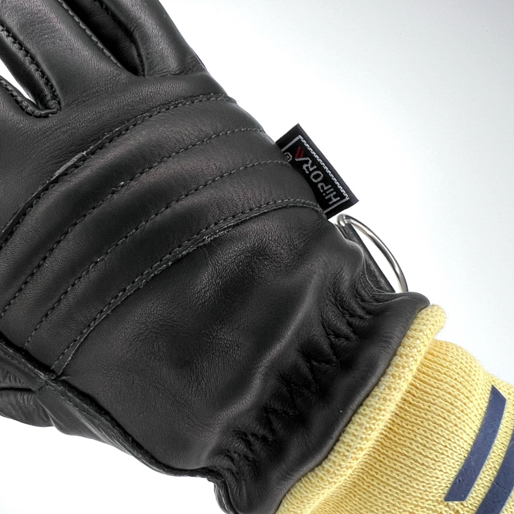 Das Bild zeigt eine Nahaufnahme der Rückhand eines schwarzen Feuerwehrhandschuhs. Deutlich zu erkennen sind die gepolsterten Verstärkungen auf dem Handrücken sowie die gelbe Manschette mit einem blauen Logo. Ein kleines Etikett mit der Aufschrift "HIPORA" ist ebenfalls sichtbar, was auf die verwendete Membran-Technologie hinweist. Der Handschuh wirkt robust und hochwertig verarbeitet.