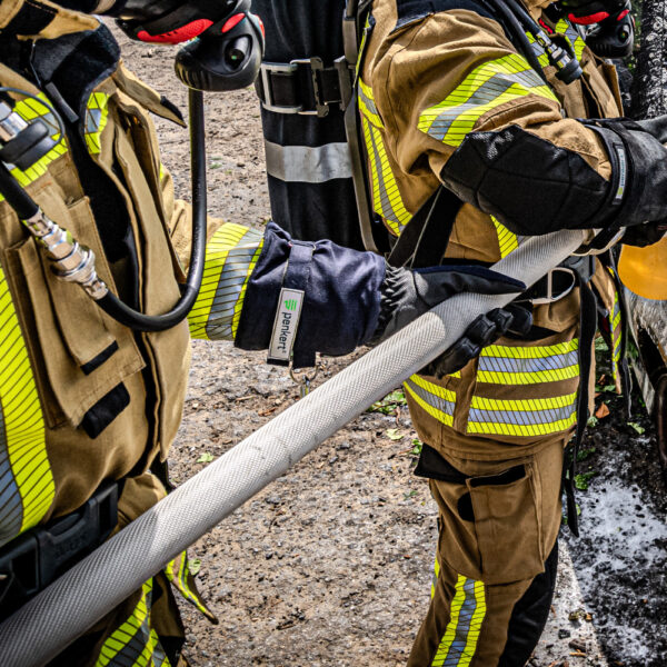 Feuerwehrkräfte in vollständiger Schutzkleidung mit reflektierenden Streifen halten gemeinsam einen Feuerwehrschlauch, während sie Schutzhandschuhe der Marke Penkert tragen.