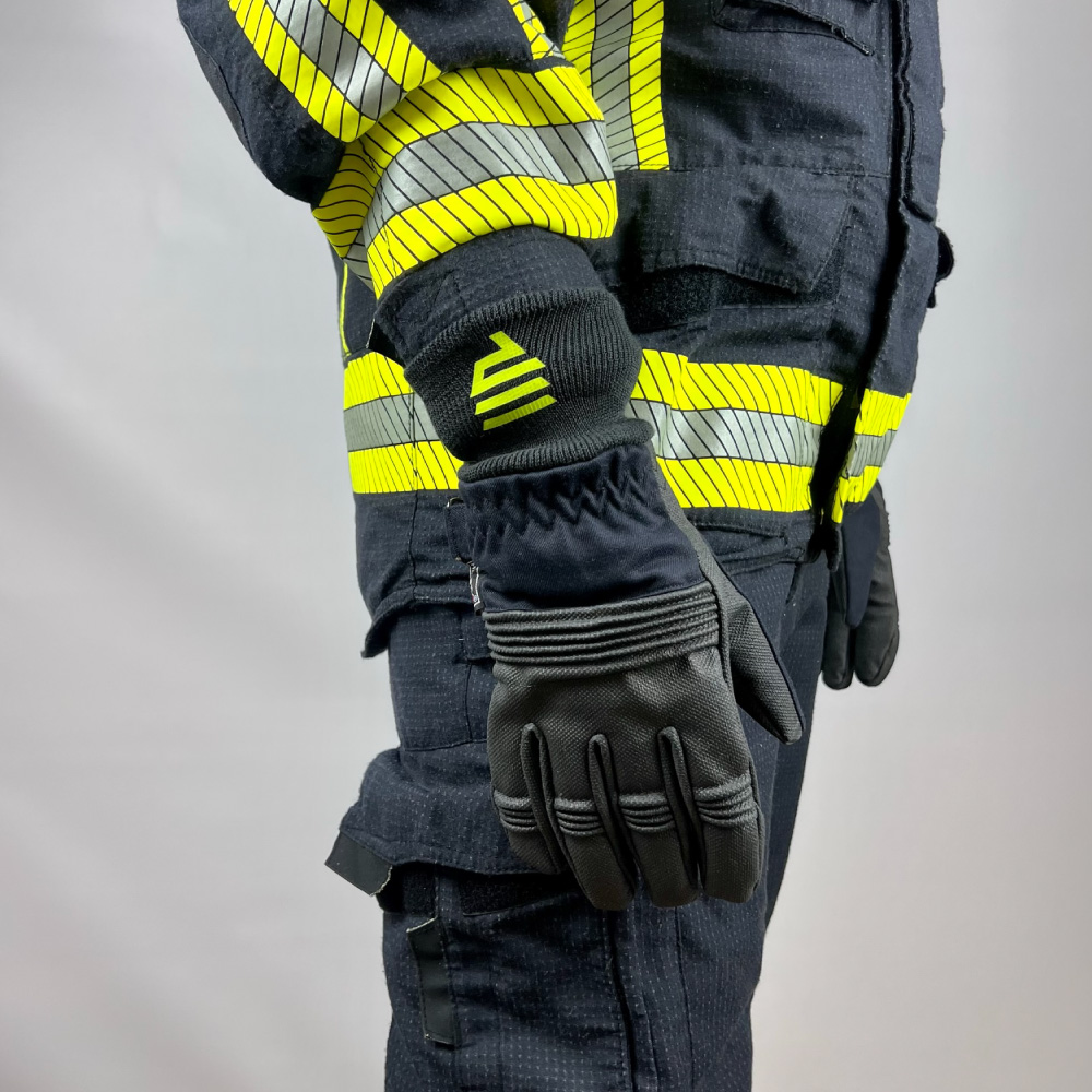 Seitenansicht einer Person in Feuerwehrschutzkleidung mit reflektierenden Streifen, die einen kurzen Schutzhandschuh mit gelbem Logo trägt.