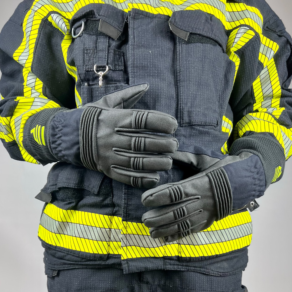 Eine Feuerwehrmann in kompletter Feuerwehrschutzkleidung trägt schwarze Schutzhandschuhe mit kurzen Stulpen und reflektierenden Streifen, die Hände übereinandergelegt.