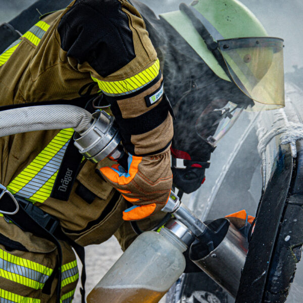 Ein Feuerwehrmann im Einsatz, der Penkert HyPA-Grip lange Handschuhe trägt und einen Schlauch an einem Feuerlöschgerät bedient, mit deutlichen orangen Fingerspitzen und robustem Design.