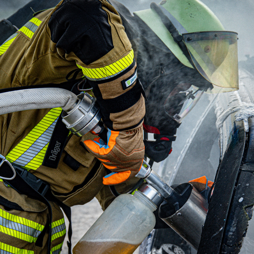 Ein Feuerwehrmann im Einsatz, der Penkert HyPA-Grip lange Handschuhe trägt und einen Schlauch an einem Feuerlöschgerät bedient, mit deutlichen orangen Fingerspitzen und robustem Design.