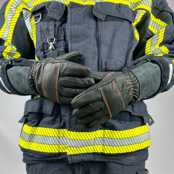 Das Bild zeigt einen Feuerwehrmann in voller Schutzkleidung mit reflektierenden gelben Streifen. Der Fokus liegt auf den schwarzen Feuerwehrhandschuhen mit langen Stulpen und orangefarbenen Nähten. Die Hände sind vor dem Körper verschränkt, wodurch die Struktur und das Design der Handschuhe sowie die Schutzkleidung deutlich sichtbar werden.