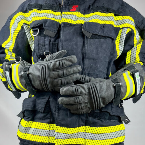 Eine Person in kompletter Feuerwehrschutzkleidung trägt lange Schutzhandschuhe mit reflektierenden gelben Streifen und Klettverschlüssen, die Hände übereinandergelegt.