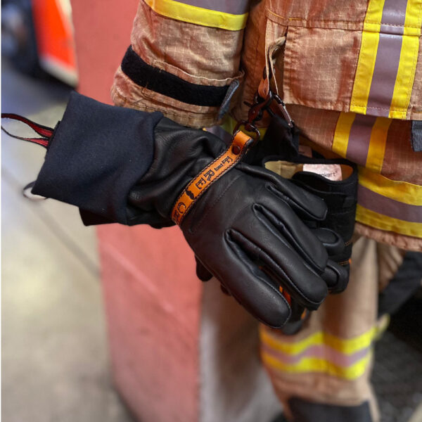 Feuerwehrmann in Einsatzkleidung mit gelben reflektierenden Streifen trägt schwarze Lederhandschuhe und hält sie mit einem Karabinerhaken an der Jacke befestigt.