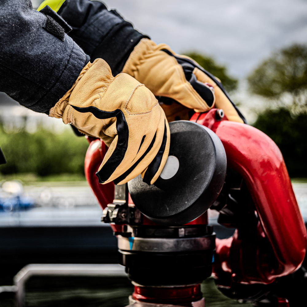 Ein Feuerwehrhandschuh Penkert Elk Defender 3.0 in Schwarz und Beige wird bei einem Einsatz verwendet, während der Träger an einem roten Feuerwehrgerät arbeitet, im Freien vor einer bewölkten Kulisse.