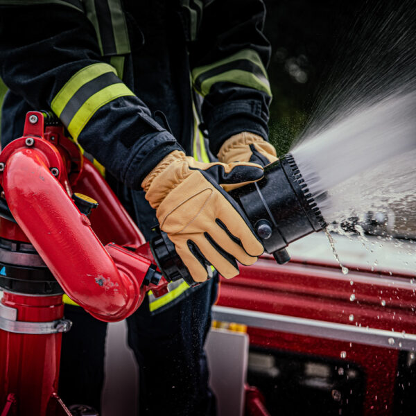 in Feuerwehrhandschuh Penkert Elk Defender 3.0 in Schwarz und Beige wird getragen, während der Träger einen Feuerwehr-Wasserwerfer bedient, aus dem Wasser spritzt